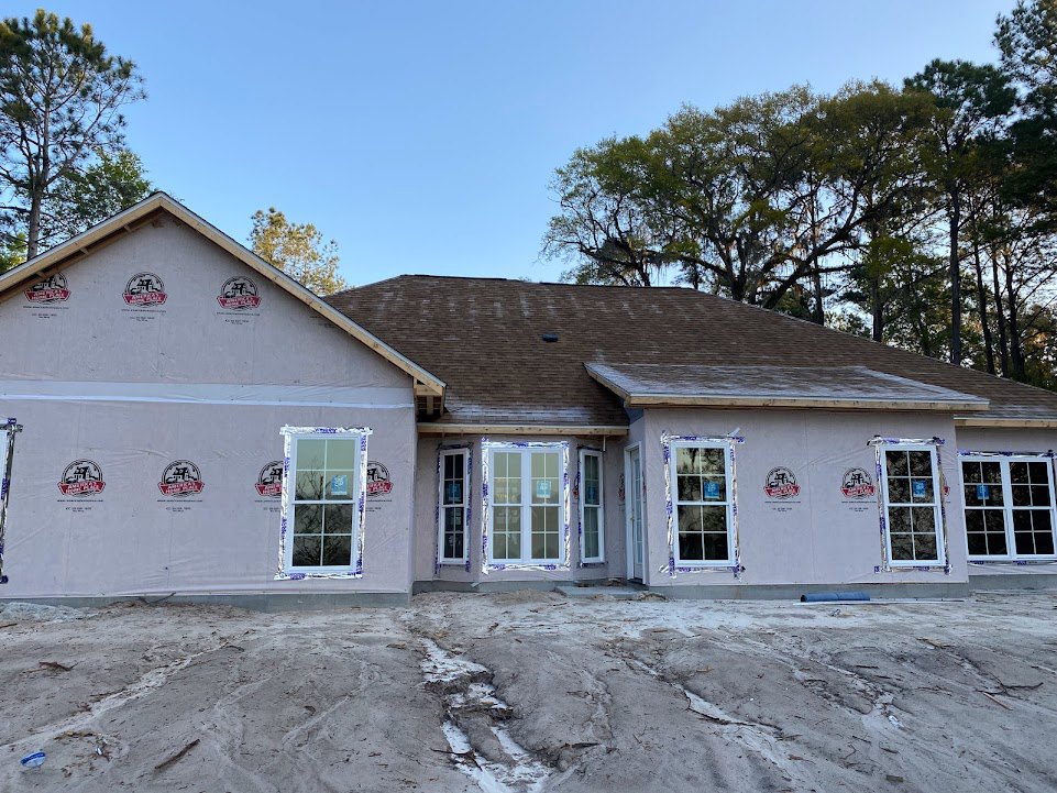 Partially built house with exposed framing, white-trimmed window, blue permit sign, shingled roof, surrounded by mature trees