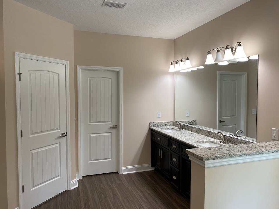 Bathroom with double sinks set in a granite countertop, large wall mirror above, wood flooring, white paneled doors with silver handles, and a row of lights mounted over the