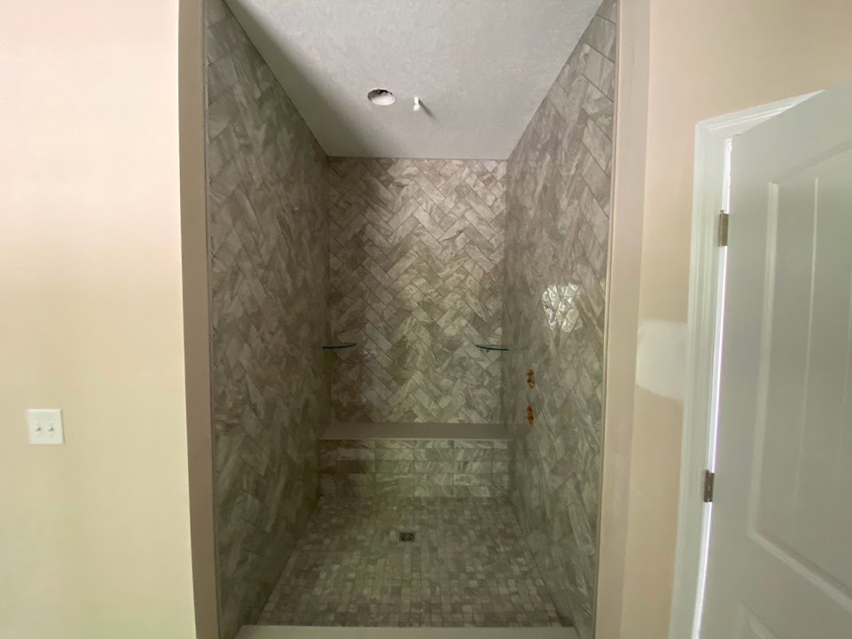 Walk-in shower with gray tile walls and floor, central drain, recessed ceiling light, and white door with matching frame.