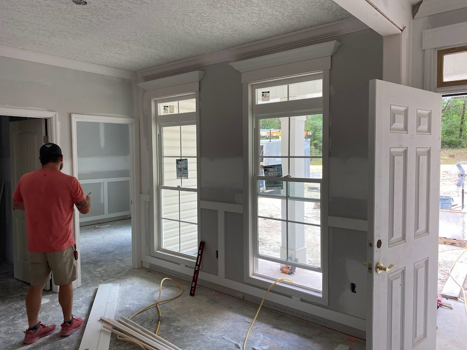 Man in red shirt and shorts standing in a bright room with white walls, large windows, wood flooring, and pink shoes nearby; window features a sign, person holding a pen.