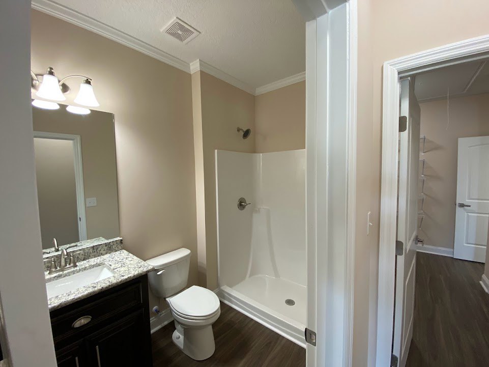 Bathroom with white toilet and sink, wall-mounted mirror above vanity, three-light fixture, white bathtub with silver handle, tiled floor, ceiling vent, close-up of brushed metal