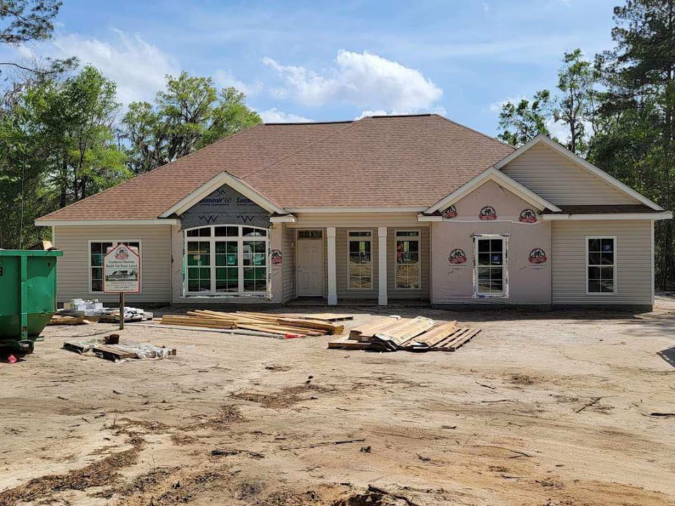 Partially built house with exposed framing, several installed windows, dirt yard, green waste container on driveway, construction sign featuring house illustration, pitched roof