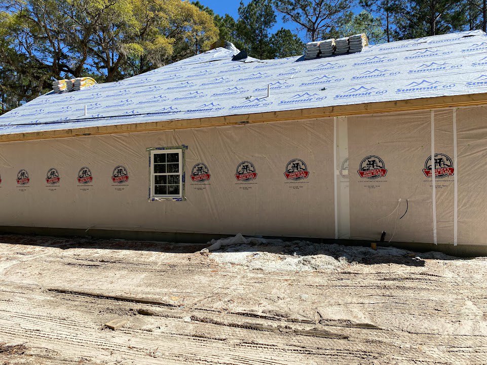 Partially built house with white sheet metal roof, exposed framing, white window frame, dirt driveway with tire tracks, surrounding trees and open sky