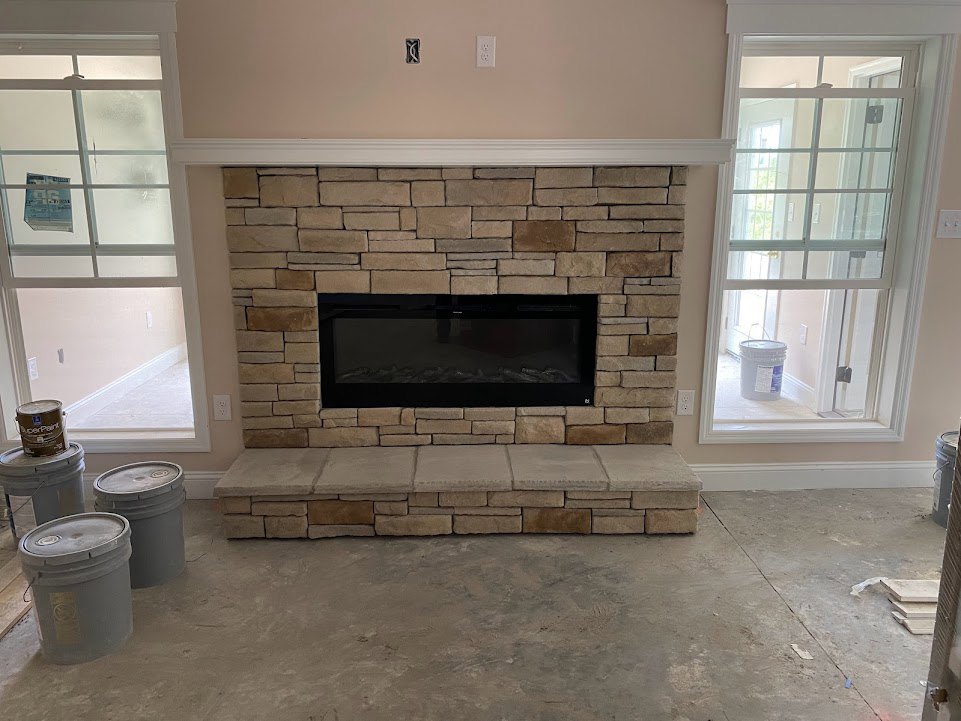 Stone fireplace with black wood-burning stove, glass-paned window, grey lidded bucket, paint can on window sill, and square-topped stone wall in den with light-colored floor.