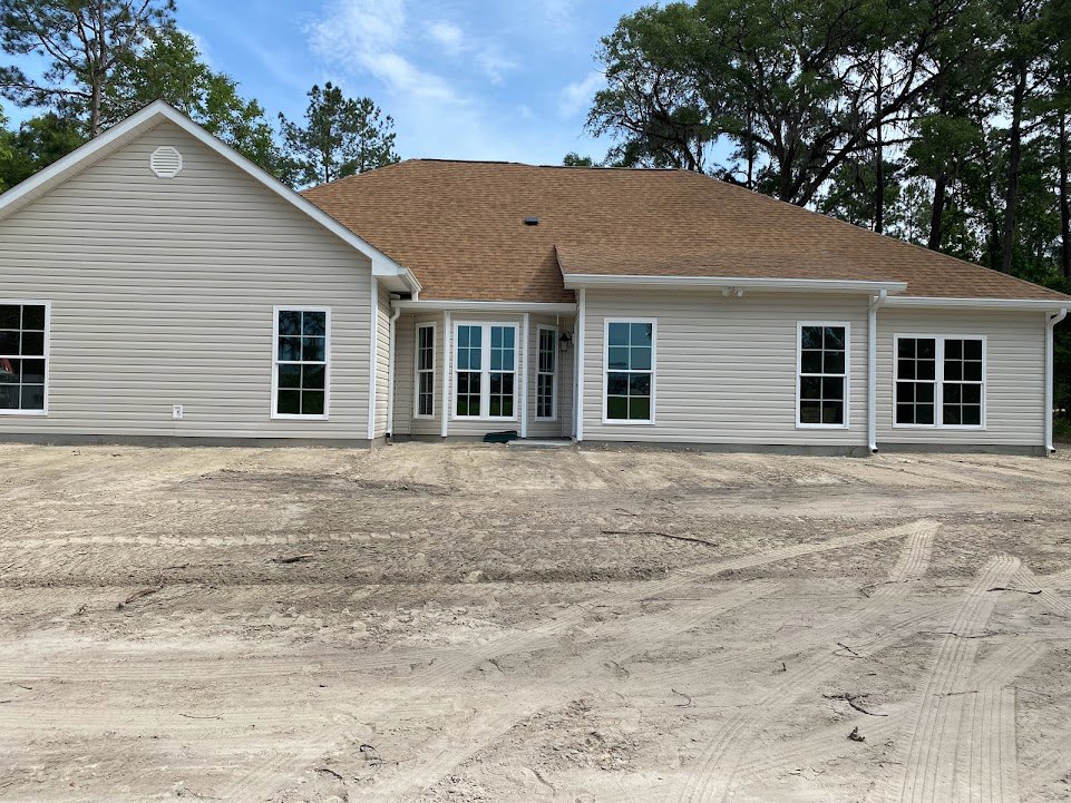 Two-story white house with brown roof, multiple white-framed windows, unfinished dirt yard, and cloudy sky in background
