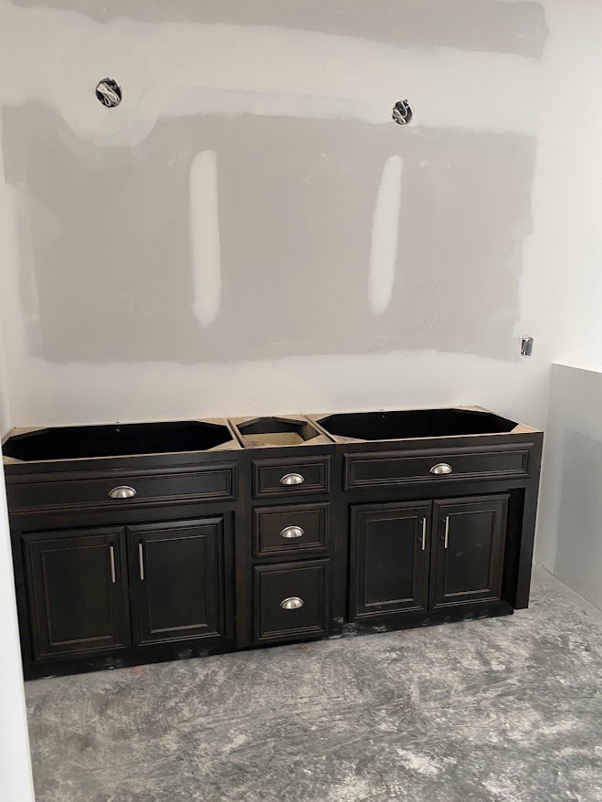 Bathroom with multiple white sinks, black cabinetry featuring silver handles, and white walls with visible holes.