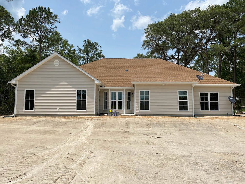 Two-story home with brown shingle roof, multi-pane windows, and exterior vents, surrounded by a dirt lot and trees under a cloudy sky