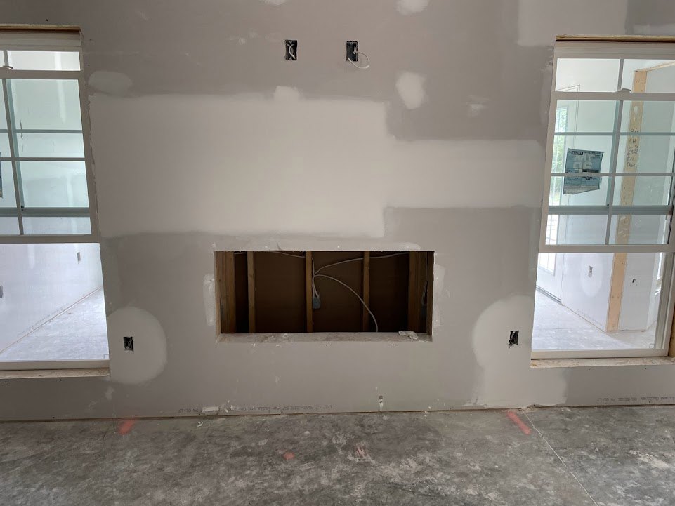 Unfinished interior with plaster wall featuring a hole and exposed white wires, concrete floor showing handprints, two windows—one with a sign, one with a glass pane—and a paper