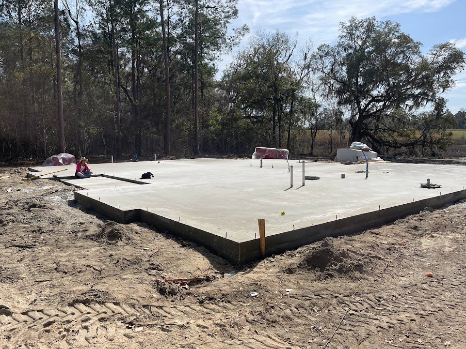 Concrete foundation slab set in a dirt field, surrounded by scattered trees under a clear blue sky, with construction poles visible along the perimeter.