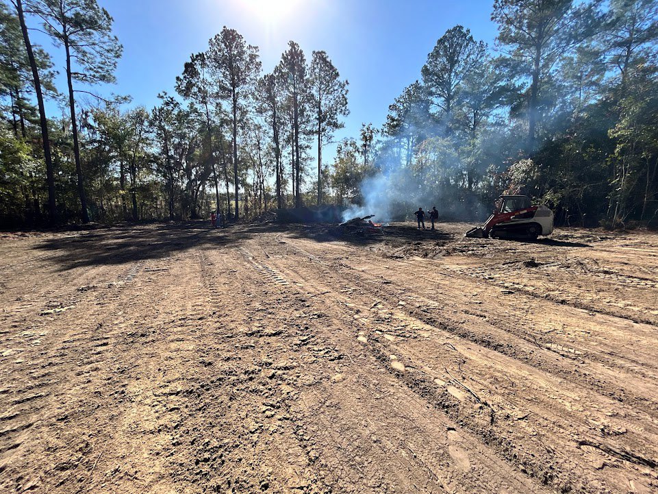 Dirt road with tire tracks bordered by tall trees, red tractor emitting smoke, group of people standing in the background, forested landscape under open sky