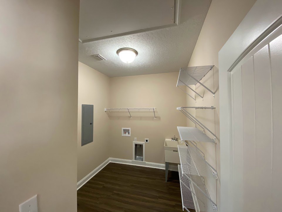 Wood floor with white baseboards, white door, grey door with square handle, white wall-mounted shelf, ceiling light fixture, white rectangular object against plaster wall