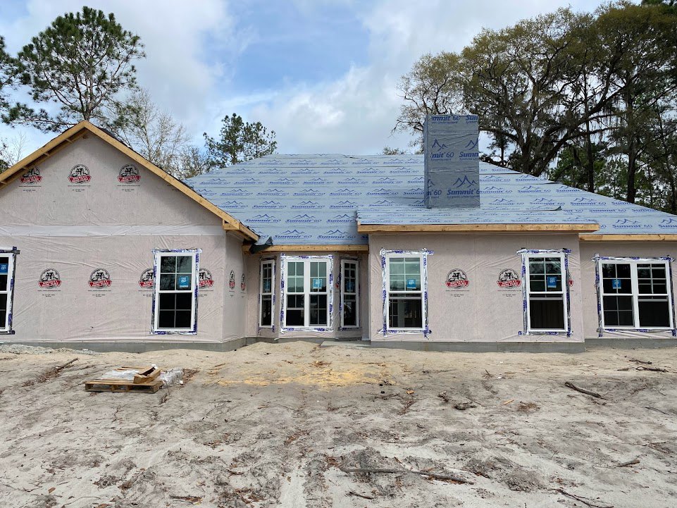 Framed house under construction with exposed plywood walls, white-framed windows, dirt and debris around foundation, grey rectangular sign with blue writing, surrounded by trees